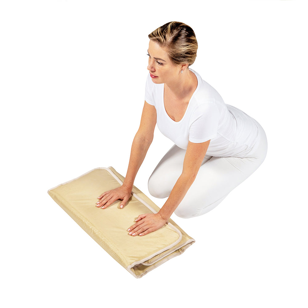 A woman in white kneels on a SOFT Mat Medium 4024 InfraMat Pro® by HealthyLine, pressing her hands on the folded amethyst mat against a white background.