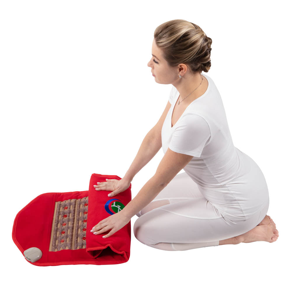 A woman in white kneels on the floor, placing her hands on a HealthyLine Photon PEMF InfraMat Pro® Amethyst Pad Medium 3618 Soft with embedded stones and control unit, set against a white background.