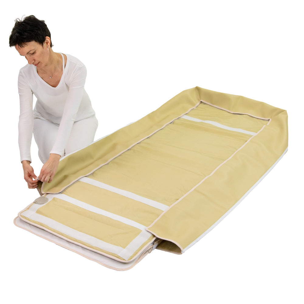 A person kneels to secure the corner of a HealthyLine 360 Wrap Set™ Platinum & SOFT Full Pro PLUS 7428 - Photon Advanced PEMF InfraMat Pro®, a beige padded heated gemstone mat with raised edges, laid flat and partly folded at one end.