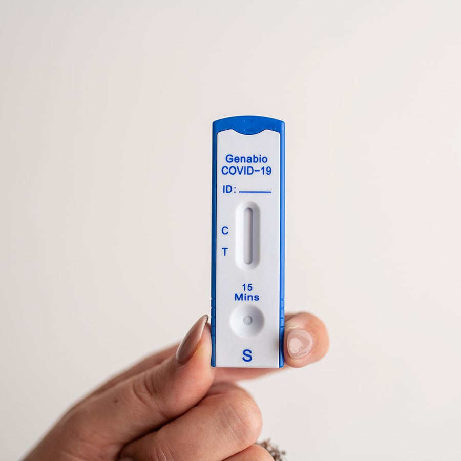 A hand holds a Unite Medical Genabio COVID-19 AT-Home Test device with a single line at “C,” indicating a negative result. The background is plain and light-colored.
