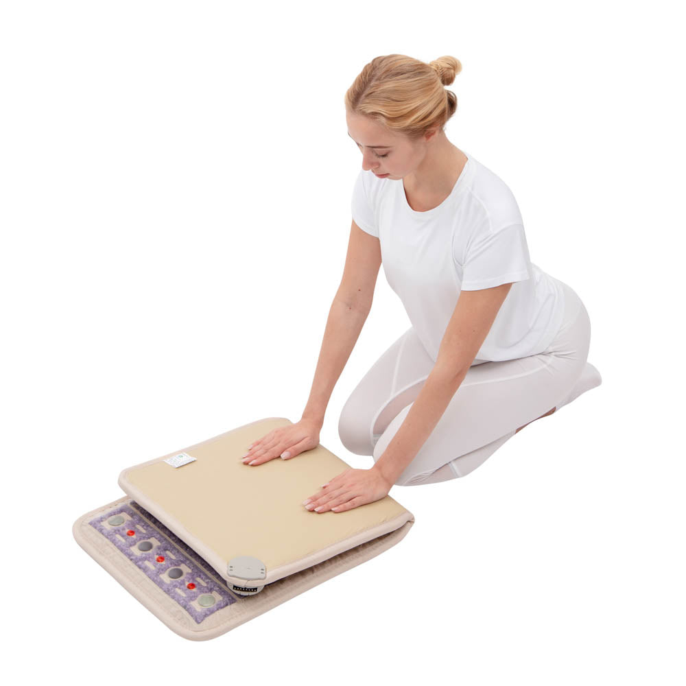 A person kneels on the floor, resting their hands on a foldable, rectangular TAJ-Mat™ Chair 4018 Firm by HealthLine, equipped with a control device. The mat's smooth surface and textured underside are adorned with colorful stones. They're dressed in white attire, enjoying natural therapies for back pain relief.