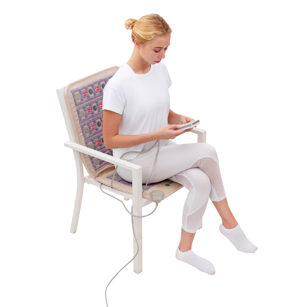 A person sits on a TAJ-Mat™ Chair 4018 Firm - Photon PEMF InfraMat Pro® by HealthLine, using a heated massage cushion with red lights designed to alleviate back pain. They hold a wired remote control. The cushion extends across the back and seat as they relax in a white outfit and socks, enjoying the combination of natural therapies.