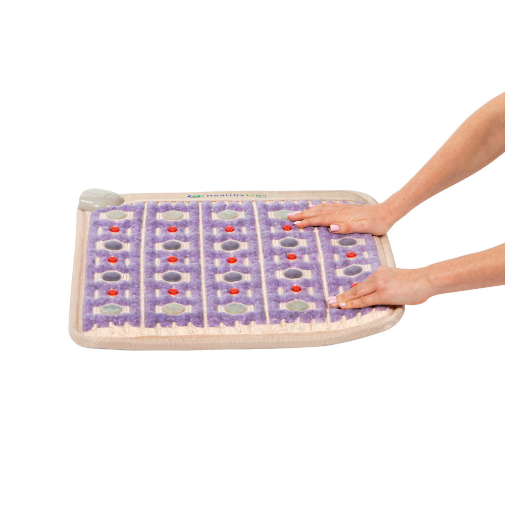 A person presses their hands on a HealthyLine TAJ-Mat™ Small 2020 Firm - Photon PEMF InfraMat Pro®, a rectangular purple infrared therapy mat with raised bumps, shown on a cream base against a plain white background.