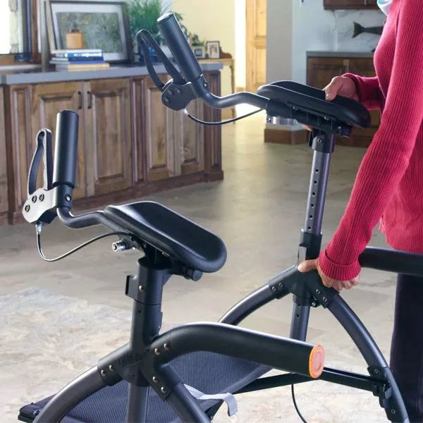 A person in a red sweater stands next to a black indoor exercise bike and a Journey Health & Life Style UpWalker H200 Standard Large in a living room with wooden cabinets and a window.