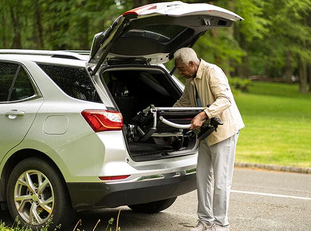 In a tranquil park, an elderly man easily loads his folded Zoomer (w/ Joystick) from Journey Health & Life Style into a white SUV's trunk, with the open rear hatch highlighting lush greenery. Dressed in a beige jacket and light pants, he's set for another seamless trip.