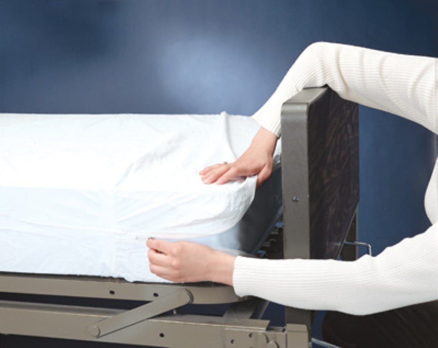 A person in a white long-sleeve shirt adjusts a fitted Graham Field Plastic Mattress Cover on a bed with a dark headboard and metallic frame. The adjustable bed, possibly for medical use, has moisture protection with PVC lining. The background is plain dark blue.