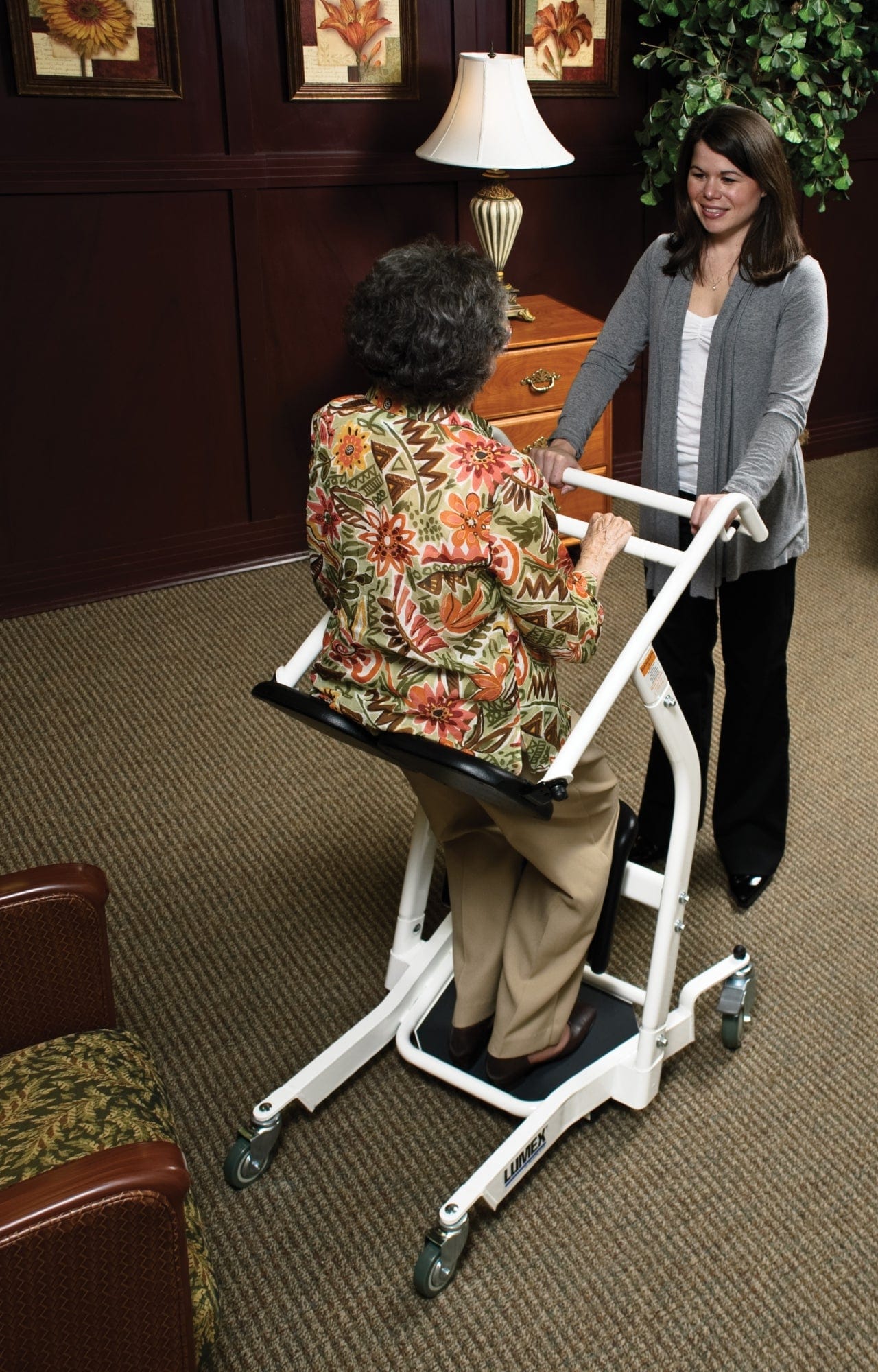 An older woman uses a Graham Field Stand Assist in a bright, carpeted room with framed art, a lamp, and wooden furniture. She faces a smiling woman offering support.