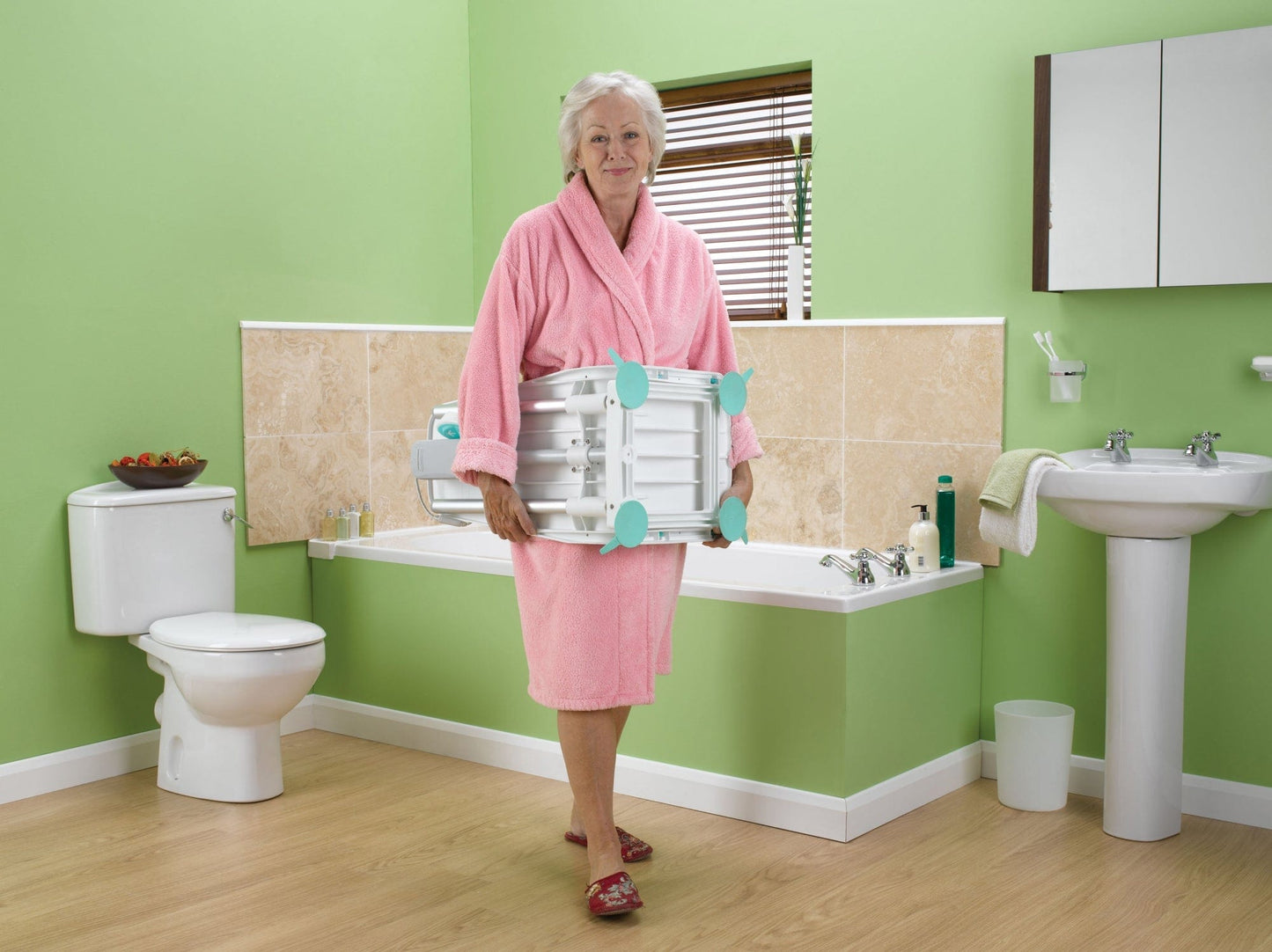 An elderly woman in a pink robe and red slippers stands in a bathroom with light green walls, beige tiles, and wooden floor, holding a Graham Field Splash Bath Lift. Around her are a toilet, sink, and toiletries on a shelf—a budget-friendly setup for comfortable bathing.