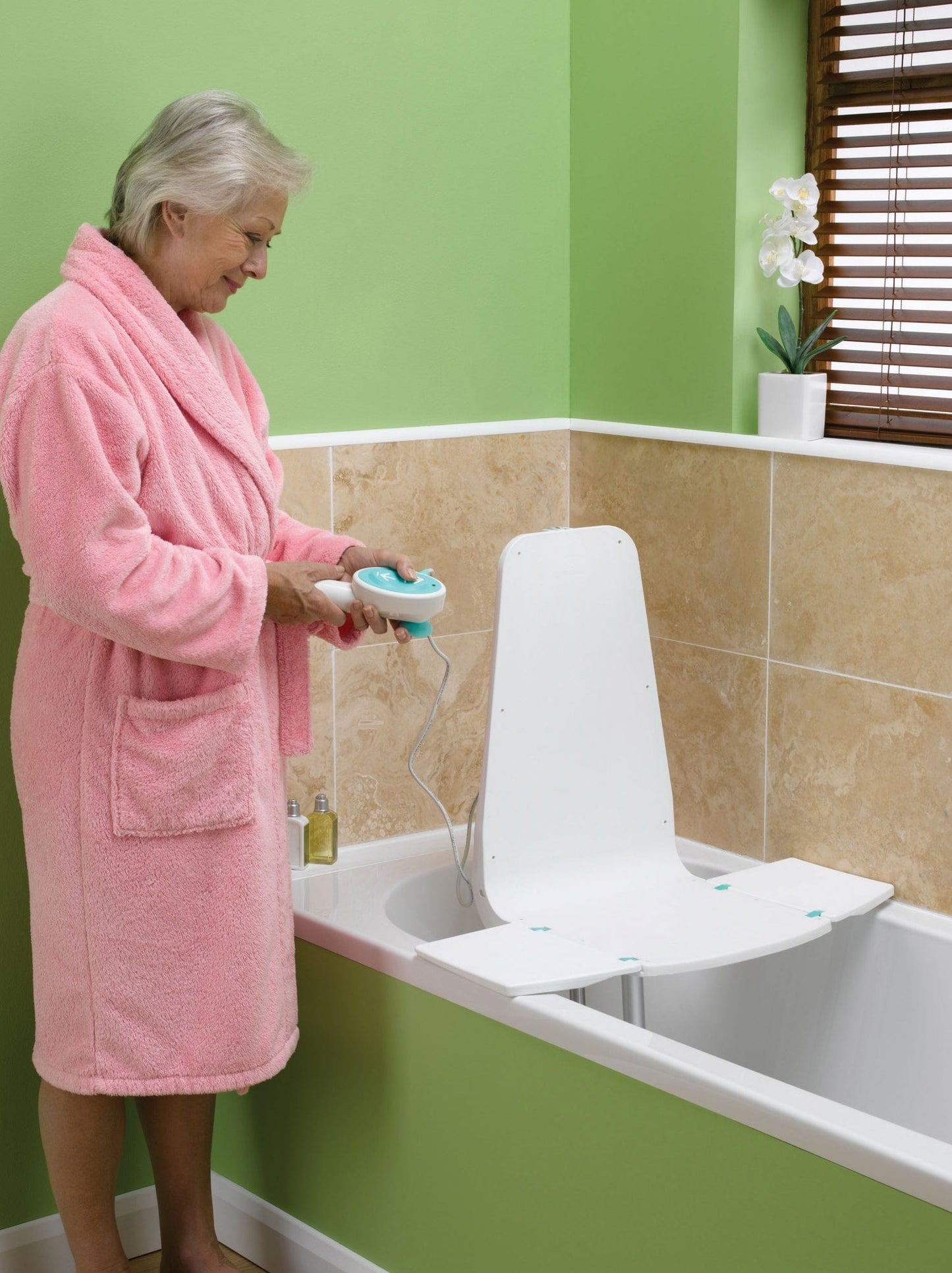 An older adult in a pink bathrobe stands next to a bathtub with the Splash Bath Lift by Graham Field. They hold a remote control, while the bathroom's green walls and beige tiles are complemented by a small plant on the windowsill. The lift is lightweight and budget-friendly.