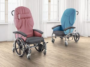 Two modern, cushioned Scoot Chair 24" TILT N Space wheelchairs from Continental Health Equipment sit in a bright room with wooden flooring and sheer white curtains. One chair is pink, the other blue; both feature adjustable seat heights, black armrests, and adjustable footrests for added comfort.