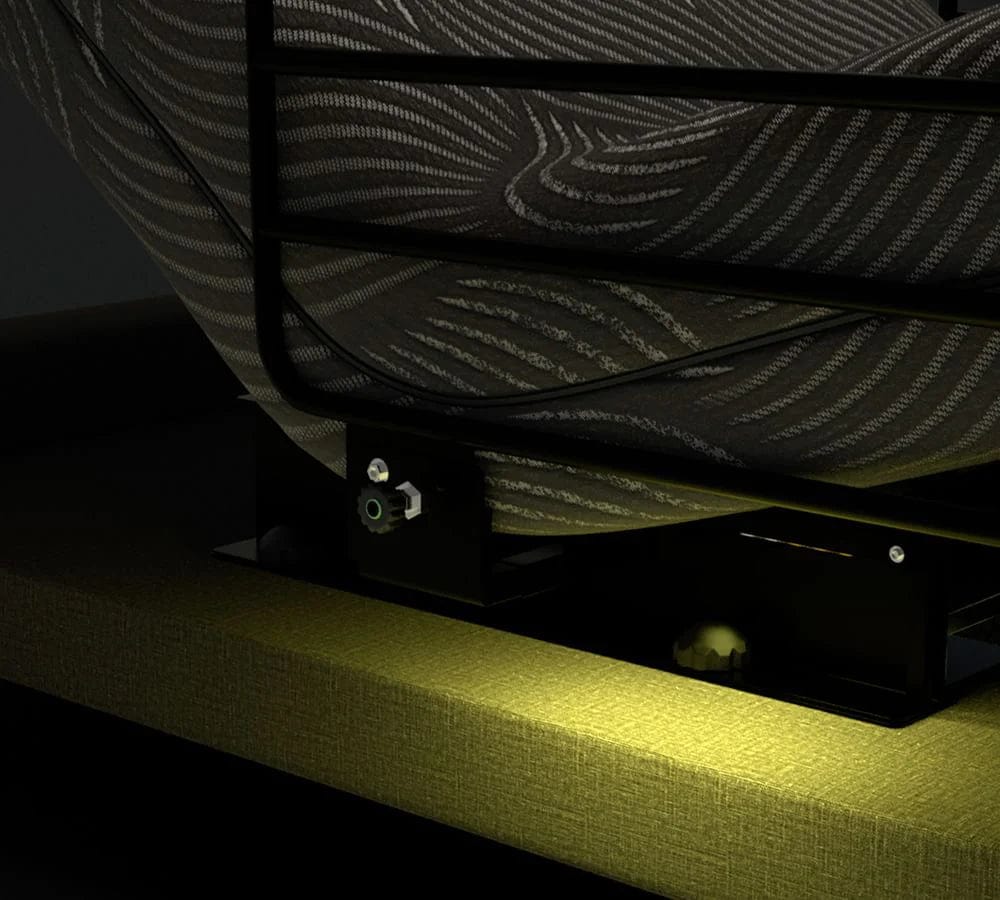 In a dimly lit scene, the base of a lift bed from Journey Health & Lifestyle's Upbed Independence is shown with a textured blanket draped over its metal frame. The adjustment mechanism of the bed is visible, casting warm light and shadows on the edge of the memory foam mattress.