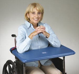 A person sits in a wheelchair, smiling warmly with hands clasped on the Skil-Care Soft Top Wheelchair Lap Tray. They are wearing a light blue shirt and beige pants, perfectly complementing the gray background. The tray features a durable vinyl cover that adds both style and functionality.