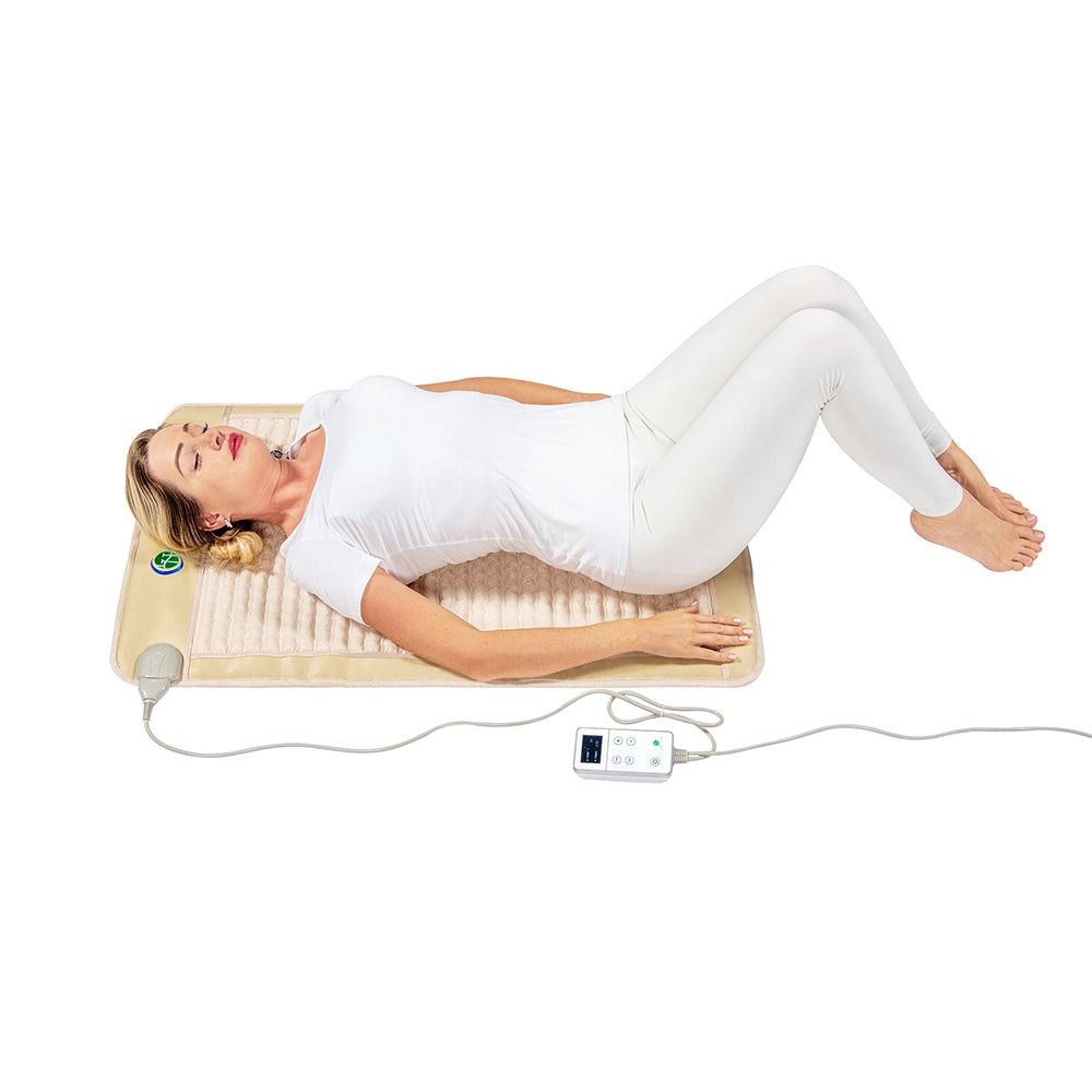 A person dressed in white is lying comfortably on the SOFT Mat Medium 4024 InfraMat Pro® by HealthyLine, a rectangular heated gemstone mat equipped with a cord and digital control panel. With knees bent and eyes closed, they are enjoying the calming warmth of the amethyst beneath them.