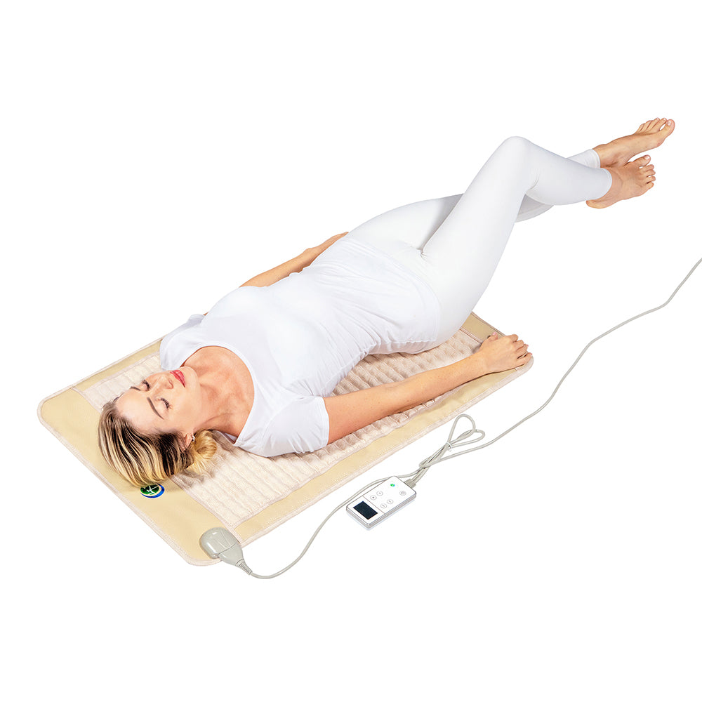 A woman in white clothing is lying on a SOFT Mat Medium 4024 InfraMat Pro® by HealthyLine, which is beige and heated with embedded amethyst gemstones. She appears relaxed, with her arms resting by her sides. The mat is connected to a remote control displaying its settings, set against a plain white background.