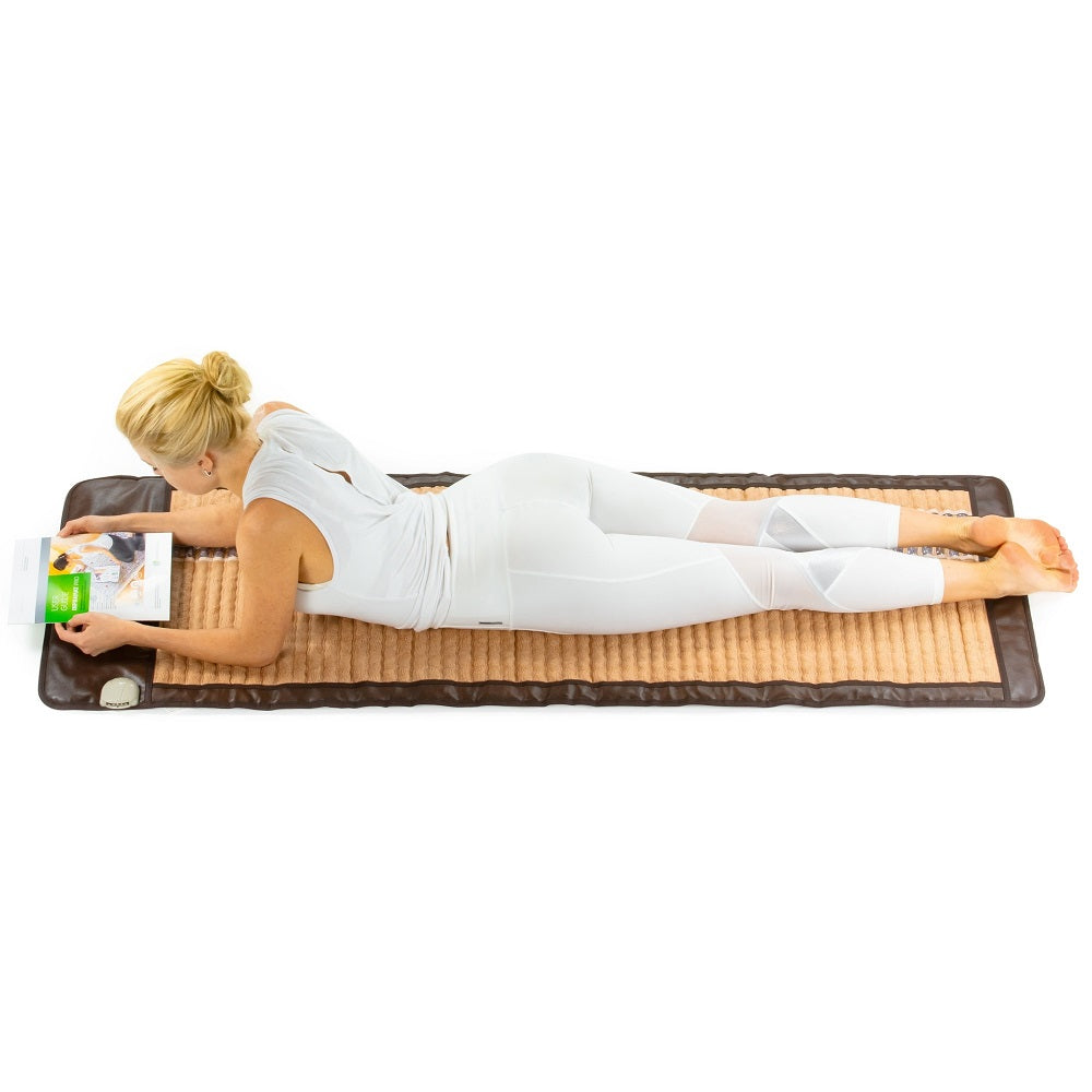 A person dressed in white clothing lies on their stomach, completely absorbed in a book, on the HealthLine SOFT-Mat™ Full 7224 InfraMat Pro®. This massage mat features a textured pattern designed for full body therapy. The individual, barefoot with short blond hair, experiences ultimate relaxation.