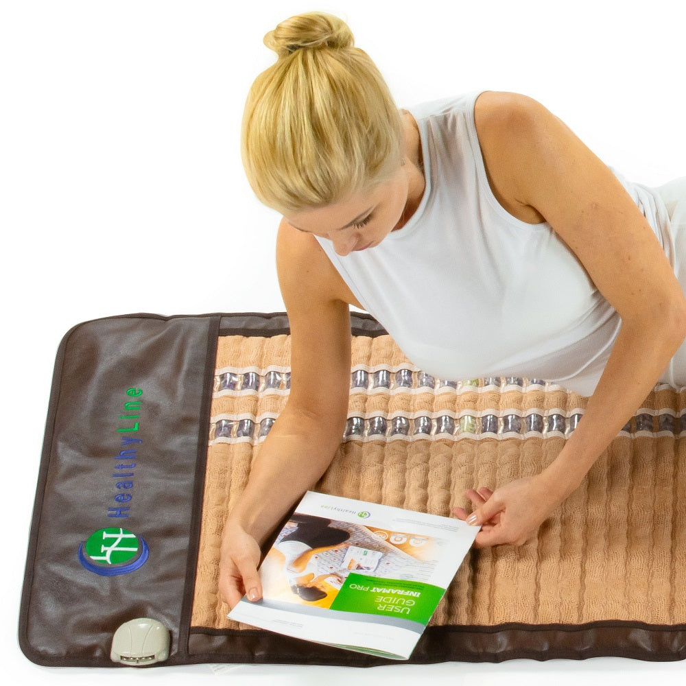 A woman with blonde hair is lying on a SOFT-Mat™ Full 7224 InfraMat Pro® from HealthLine, reading a brochure. The mat, adorned with the HealthLine logo, features rows of embedded stones designed for full body therapy. She is dressed in a white sleeveless top.
