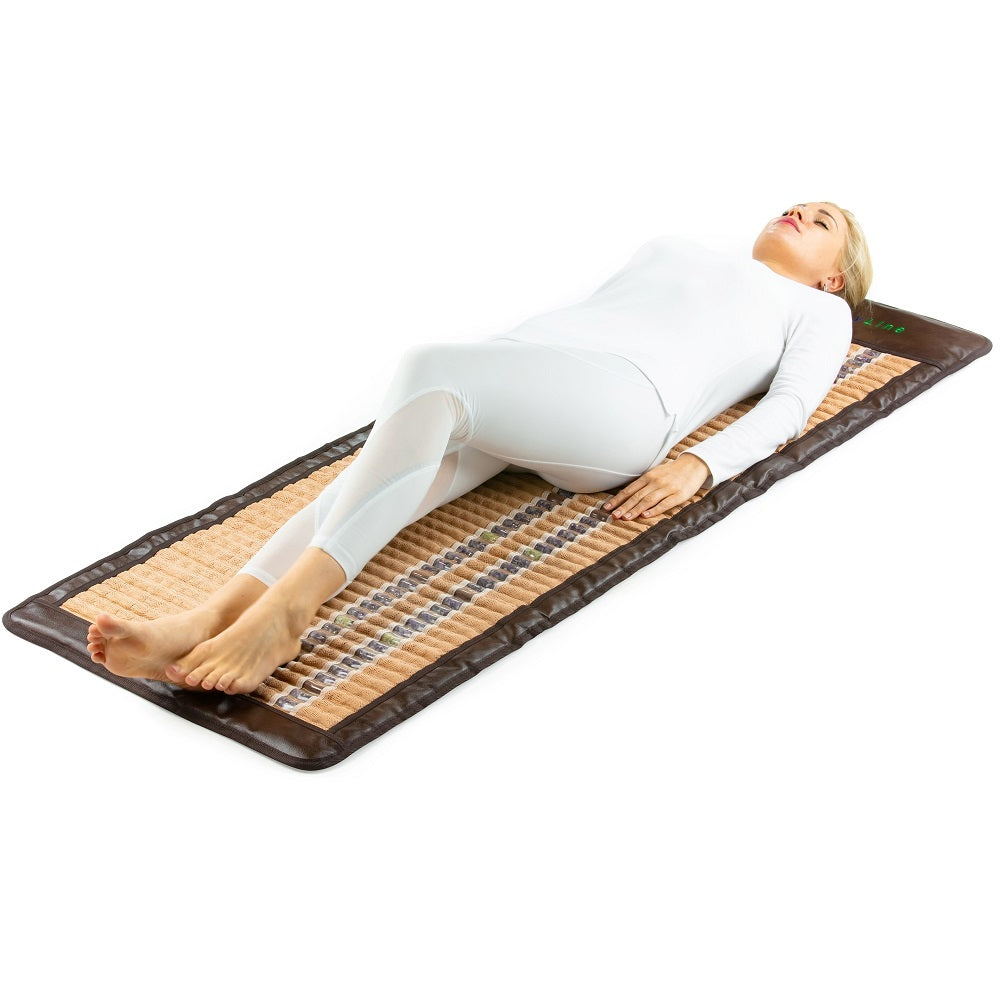 Dressed in white, a person is reclining on the HealthLine SOFT-Mat™ Full 7224 InfraMat Pro®, a mat with a specially designed layered surface for comprehensive body therapy. The gemstone composition of the mat further enhances comfort and relaxation. A plain white background highlights both the individual and the mat.