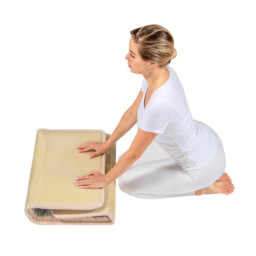 A person in a white outfit kneels to touch a folded beige mattress resembling the Rainbow Chakra Mat™, Small 4020 Firm by HealthyLine, on the floor against a minimalistic white background.