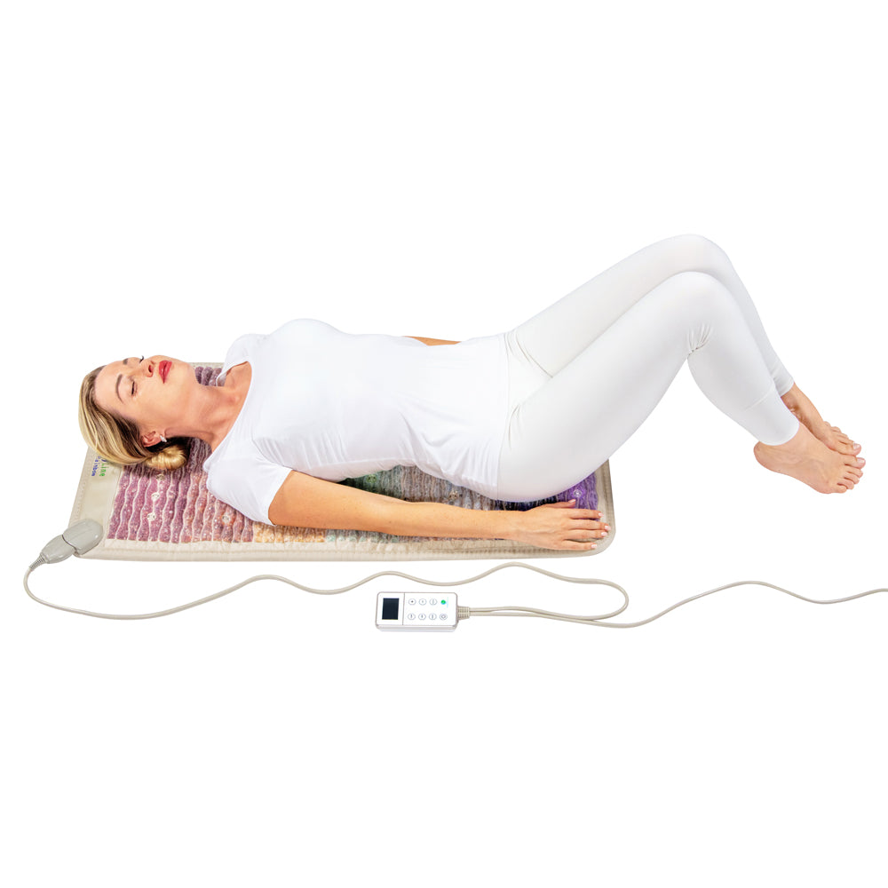 A woman in a white outfit is lying on a Rainbow Chakra Mat™ Small 4020 Firm - Photon PEMF Inframat Pro® 3rd Edition by HealthyLine with her knees bent and eyes closed. A control device connects to the mat, which emits far-infrared rays for enhanced relaxation, against a plain white background.