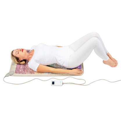 A woman in a white outfit is lying on a Rainbow Chakra Mat™ Small 4020 Firm - Photon PEMF Inframat Pro® 3rd Edition by HealthyLine with her knees bent and eyes closed. A control device connects to the mat, which emits far-infrared rays for enhanced relaxation, against a plain white background.
