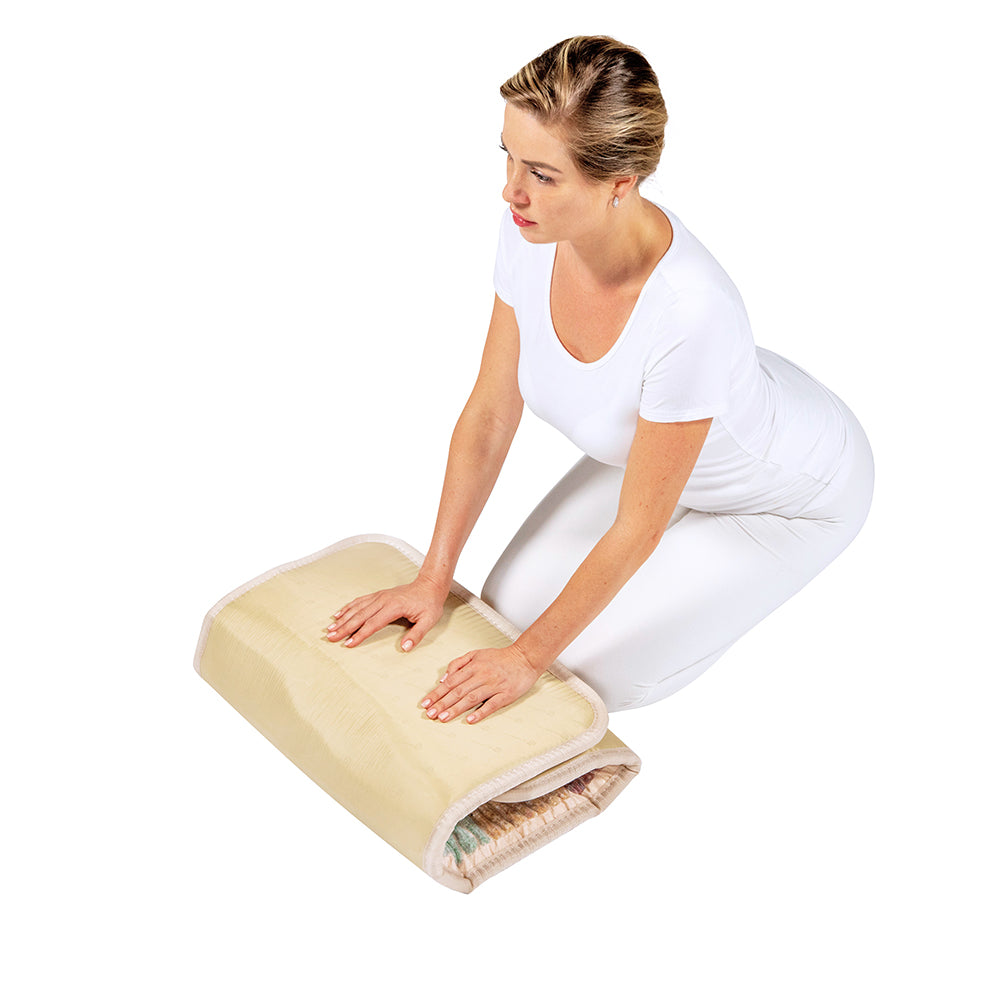 A person in a white outfit kneels on the floor, pressing down on a folded beige bed sheet atop a HealthyLine Rainbow Chakra Mat™ Small 4020 Firm - Photon PEMF Inframat Pro® 3rd Edition against a plain white background.