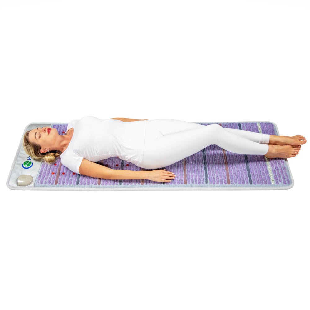 A person lies on a Platinum Mat™ Full 7224 Firm by HealthyLine, their eyes closed, dressed in a white outfit. The mat's textured surface and attached control device are specifically designed for gemstone heat therapy. The scene is set against a plain white background.