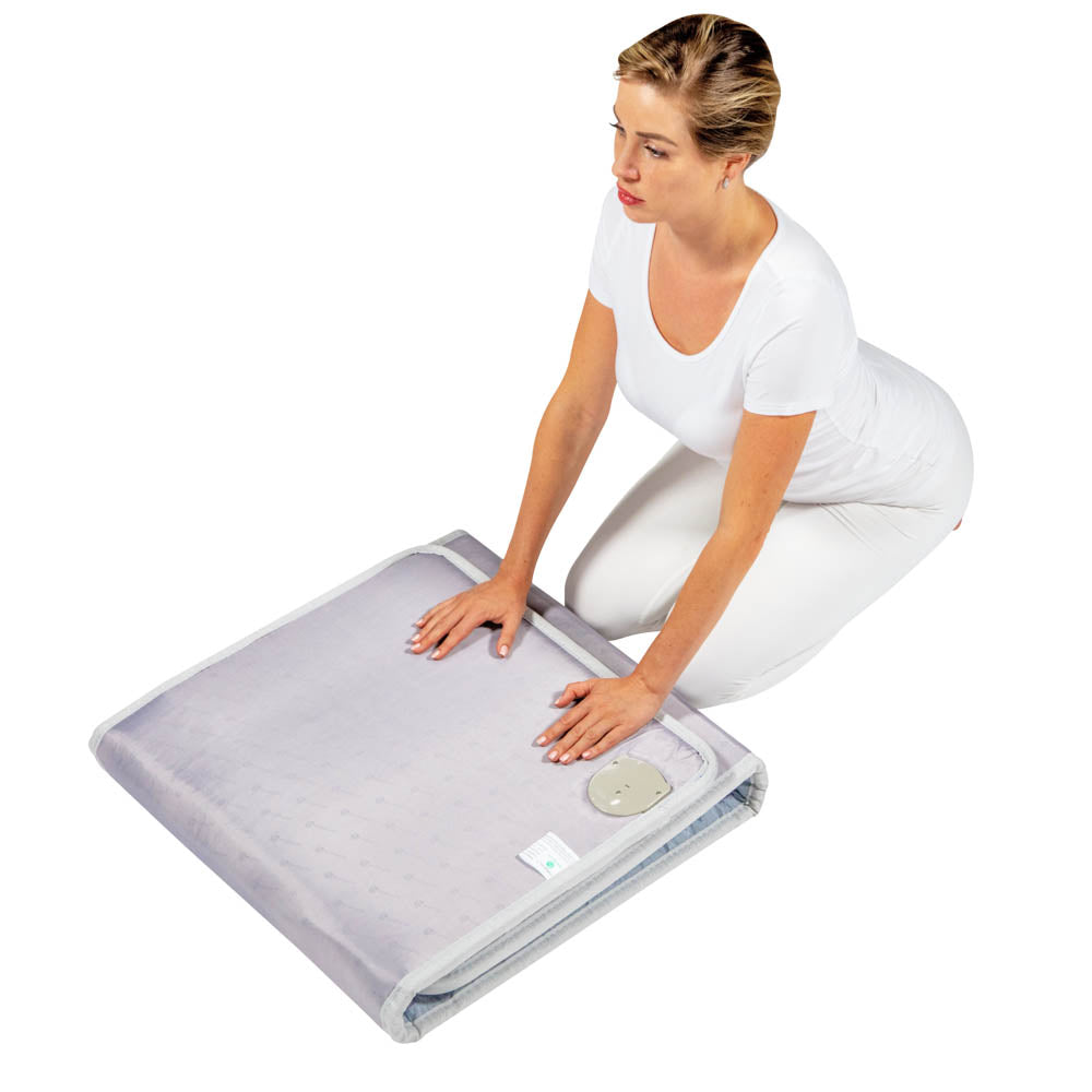 Dressed in white, a person kneels on the floor, carefully folding a large, light-gray Platinum Mat™ Full 7224 Firm from HealthyLine. This versatile Photon Advanced PEMF InfraMat Pro® could be equipped with features such as a PEMF system or gemstone heat therapy, all set against a plain white background.