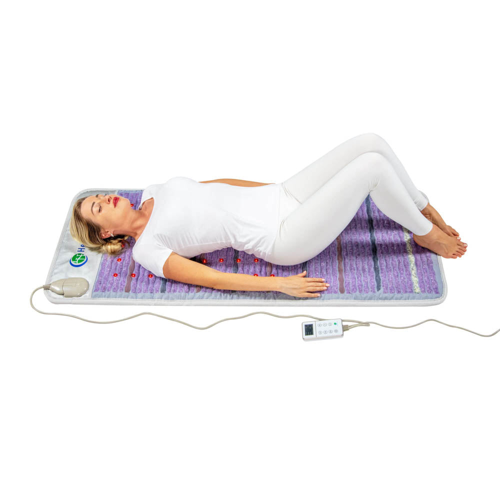 A woman in white lies on her back on the HealthyLine Platinum Mat Full Short 6024 with 30 Photon LED and advanced PEMF, knees bent, eyes closed, hands by her sides. The mat’s customizable PEMF remote is attached by a cord.