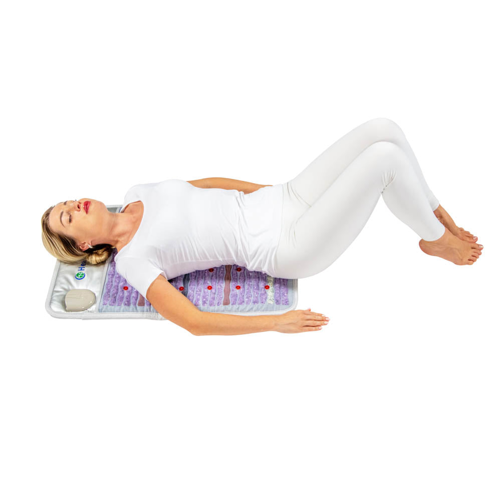 A woman in a white outfit lies on her back on the HealthyLine Platinum Mat™ Medium 3220 Firm - Photon Advanced PEMF InfraMat Pro®, featuring various textures and colors. Photon lights enhance her relaxation, while the plain white background complements the serene scene. Her hands rest peacefully by her side, immersed in tranquility.