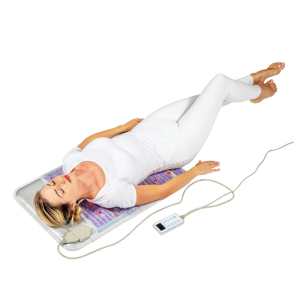 Clad in white, a person reclines with closed eyes and arms at their sides on the HealthyLine Platinum Mat™ Medium 3220 Firm - Photon Advanced PEMF InfraMat Pro®. The textured surface features visible round elements, while soothing photon lights intensify the relaxation experience as they hold a remote control.
