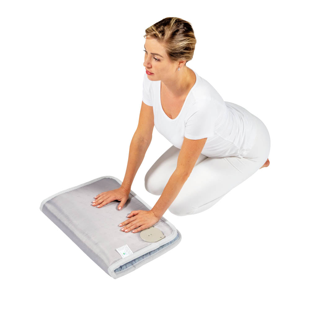 A person kneels on the floor, pressing their hands onto a gray Platinum Mat™ Medium 3220 Firm - Photon Advanced PEMF InfraMat Pro® by HealthyLine. They are dressed in white clothing against a plain white background, enhancing the serene atmosphere.