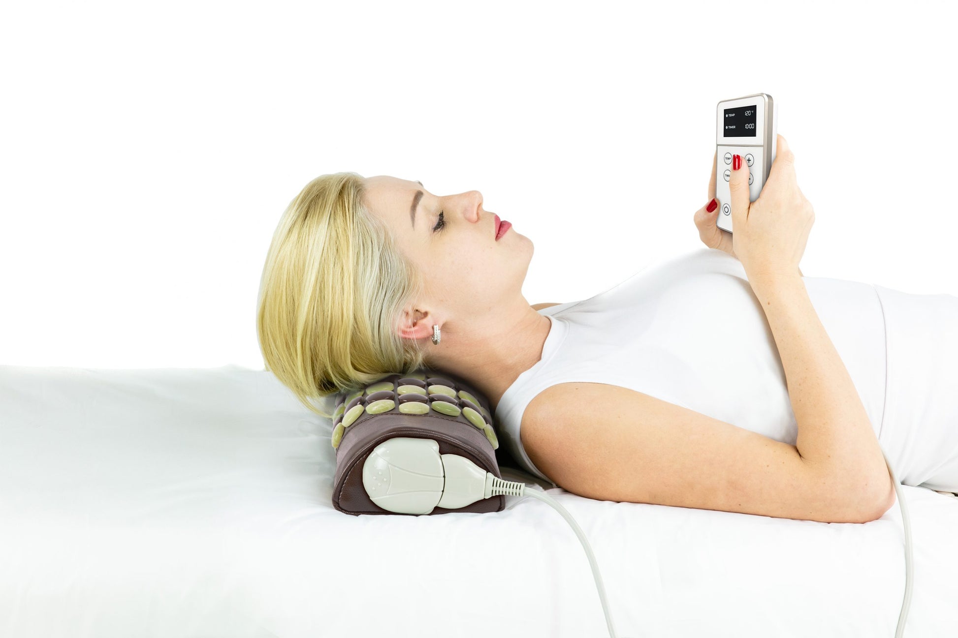 A woman with blonde hair is lying on a Pebble JT Bolster Firm InfraMat Pro® by HealthyLine, resting her head on a specialized pillow. She is holding and examining a handheld remote device. The setting is bright, with a white background, enhanced by the far-infrared rays emitted by jade and tourmaline gemstones.