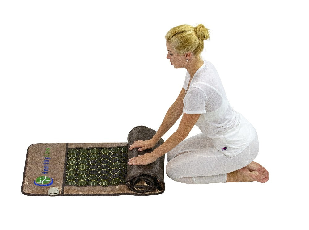A person dressed in white kneels on the floor, meticulously rolling up a HealthyLine Mesh JT Mat Full 7224 Flexible InfraMat Pro®. This brown mat, adorned with circular patterns and infused with jade and tourmaline, includes a control device and features a logo displaying a globe and leaf design, making it perfect for far infrared therapy.