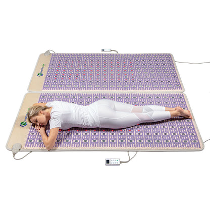 A person in white athletic clothing is reclining on their side on two large TAJ-Mat™ King 8076 Firm - Photon PEMF Split Inframat Pro® heating mats by HealthyLine, which are connected to a control device. They are enjoying the benefits of PEMF therapy in a bright and minimalistic setting.