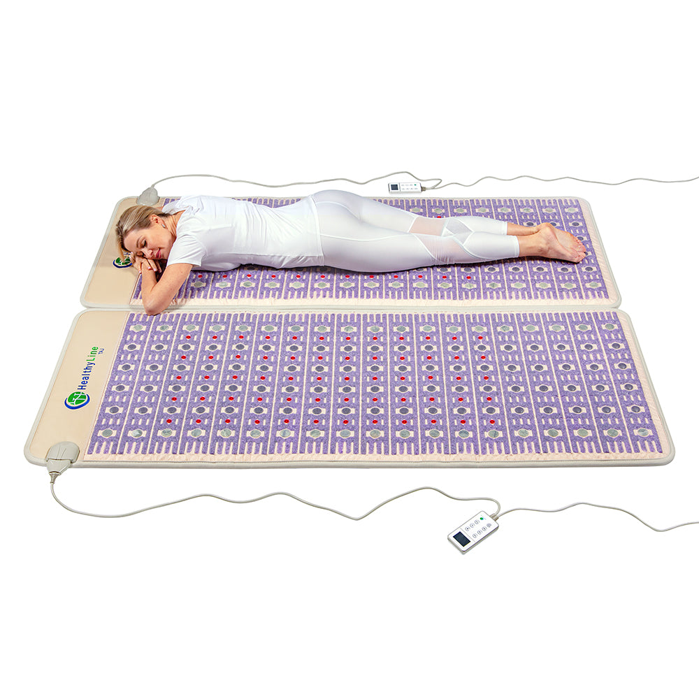 A person lies face down on a large purple TAJ-Mat™ Queen 8060 Firm - Photon PEMF Split Inframat Pro® by HealthyLine, wearing white. Geometric patterns and two remote controls for far infrared heat adjustments are visible, indicating soothing PEMF therapy capabilities.