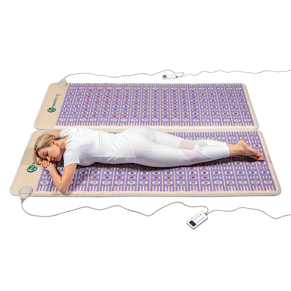 A person in white clothing lies peacefully on a TAJ-Mat™ Queen 8060 Firm by HealthyLine, enveloped in far infrared heat. This large mat includes soft purple and pink accents with a remote control for easy adjustments, creating an ideal space for relaxing and unwinding.