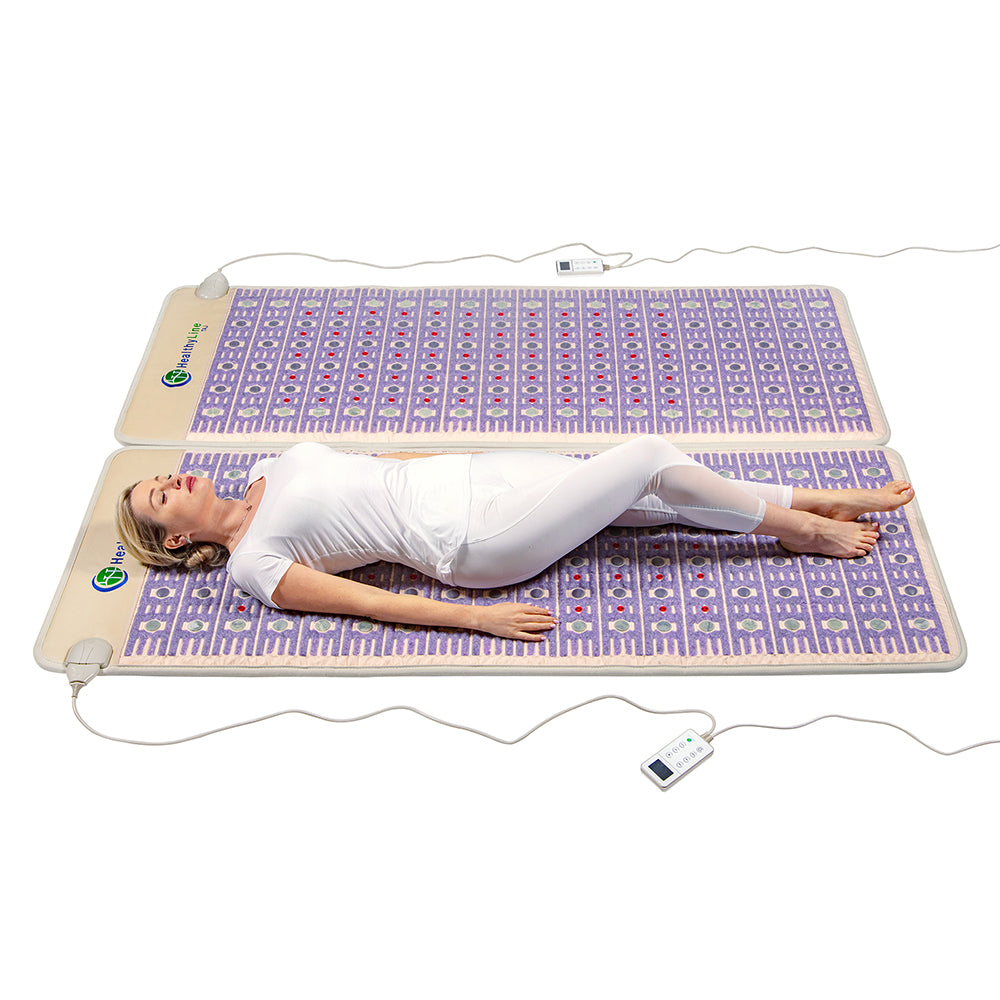 A person dressed in white lounges on a TAJ-Mat™ Queen 8060 Firm from HealthyLine, an expansive, rectangular mat with purple patterns. This Photon PEMF Split Inframat Pro® provides therapeutic relaxation using far infrared heat. A white controller is attached, and it lies on a comfort-enhancing white surface.