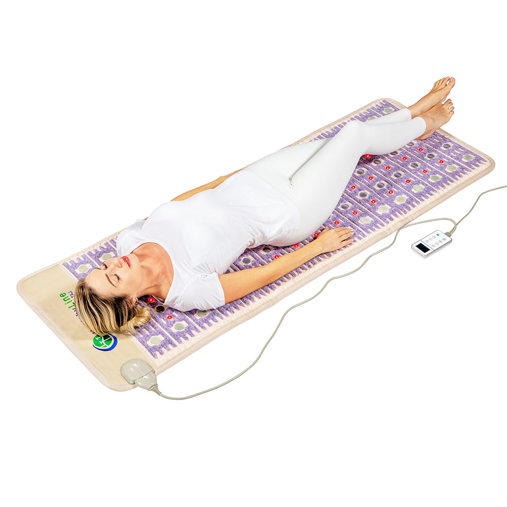 A person in white clothing relaxes on the TAJ-Mat™ Full 7224 Firm - Photon PEMF InfraMat Pro® by HealthyLine, adorned with a purple pattern. Wired to a control device, it enhances wellness. The serene white background and their closed eyes emphasize tranquility.
