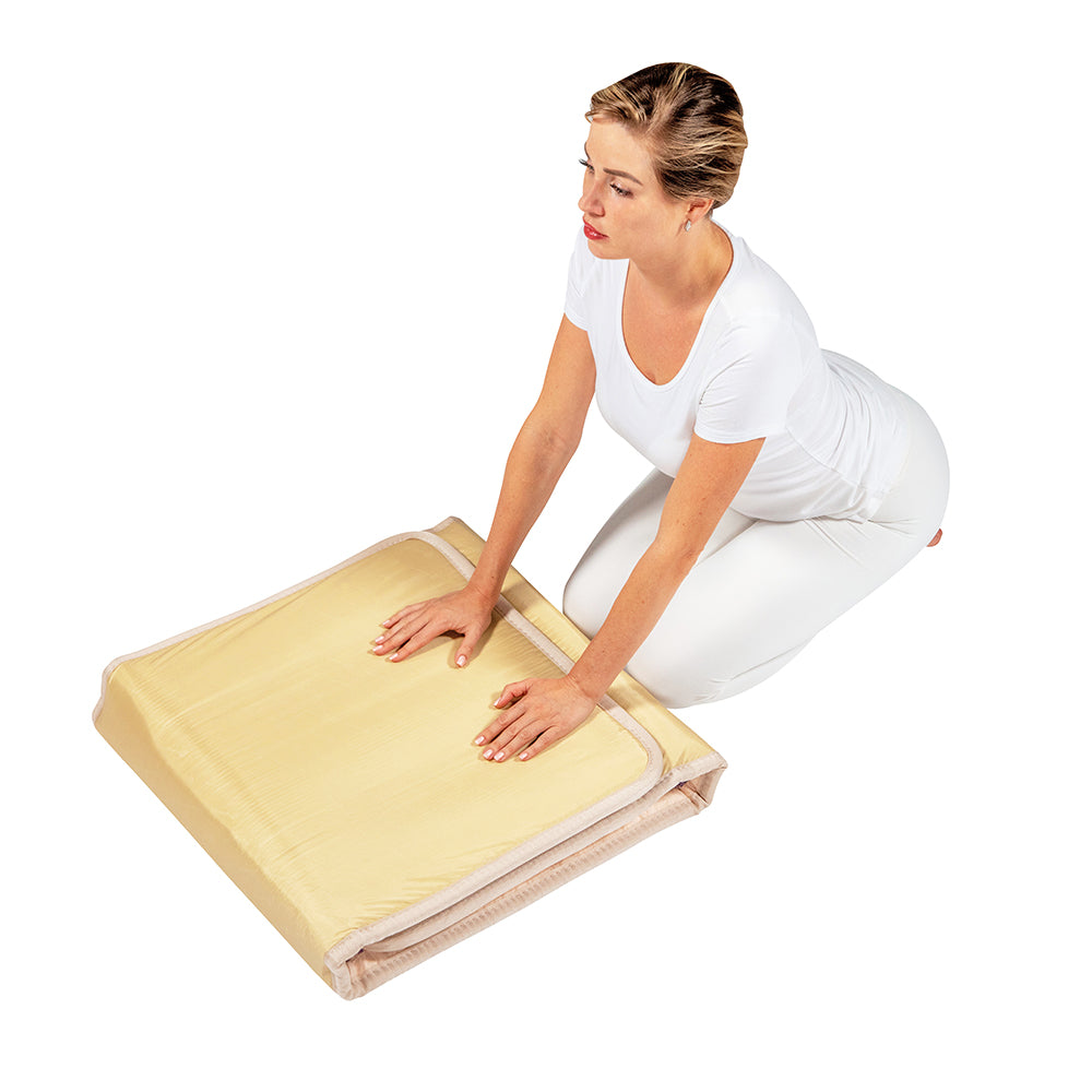 A person kneels on the floor, pressing down with both hands on a folded, light-colored foam product. Dressed in white against a plain backdrop, they enjoy wellness benefits from the luxurious TAJ-Mat™ Full 7224 Firm - Photon PEMF InfraMat Pro® by HealthyLine.