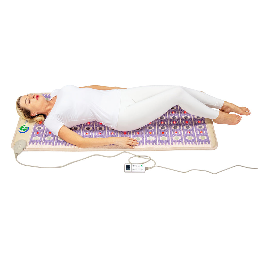 Someone in white clothing is lying on the HealthyLine TAJ-Mat™ Full Short 6024 Firm - Photon PEMF InfraMat Pro®, a purple, patterned heating mat for full-body therapy. It's connected via a cable to a control device, delivering natural therapies that relax the individual with closed eyes and slightly bent legs.