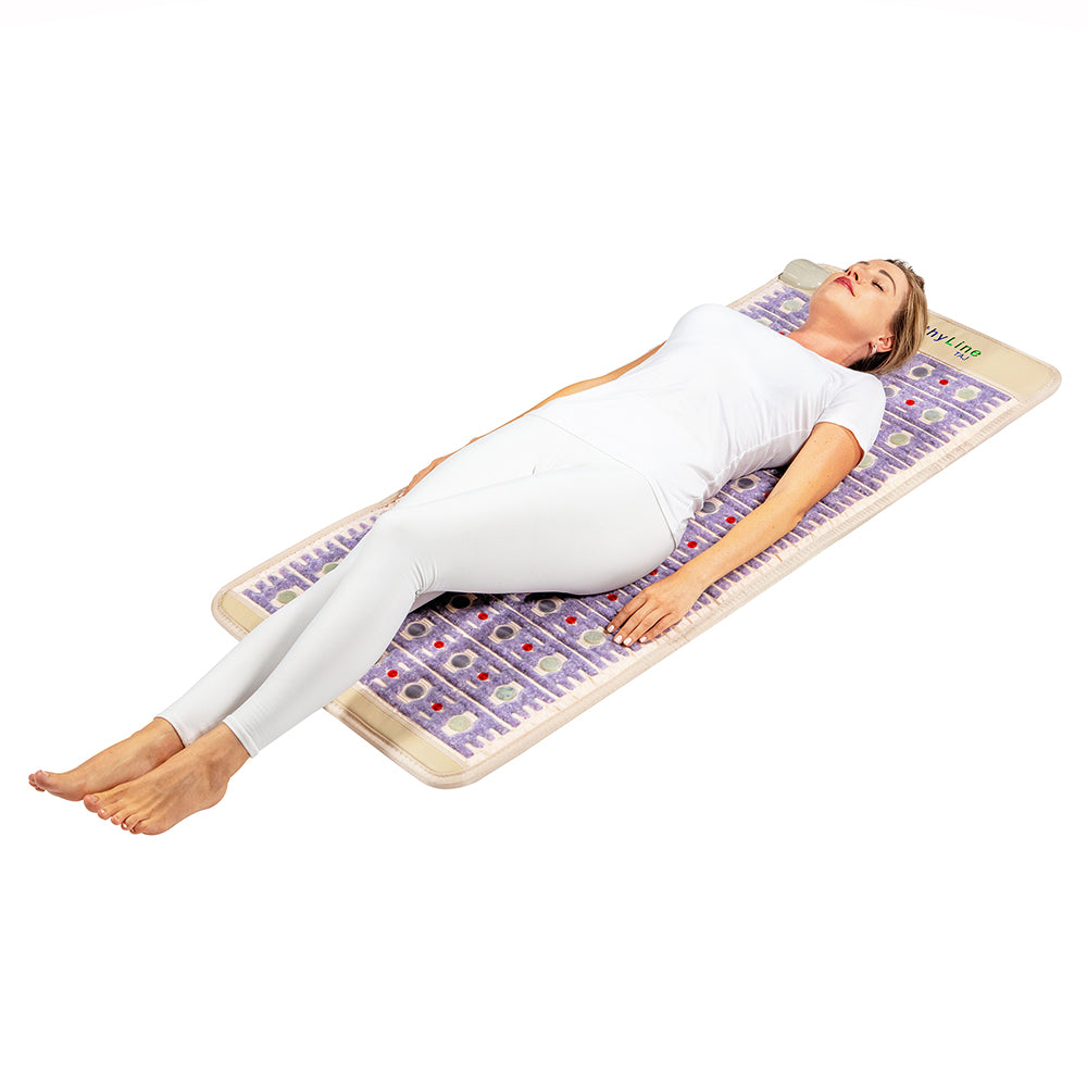 Clad in white, a person lies on the TAJ-Mat™ Full Short 6024 Firm - Photon PEMF InfraMat Pro® by HealthyLine, which showcases purple and beige circular designs. They rest with eyes closed, hands by their sides, embodying serene relaxation through natural therapies against a calm white backdrop.