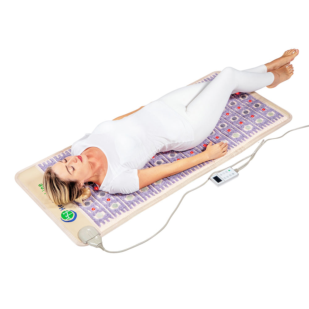 A person in a white outfit lies on the TAJ-Mat™ Full Short 6024 Firm - Photon PEMF InfraMat Pro® by HealthyLine, a therapeutic cream mat with embedded stones and a digital control panel, perfect for relaxation or heat therapy.