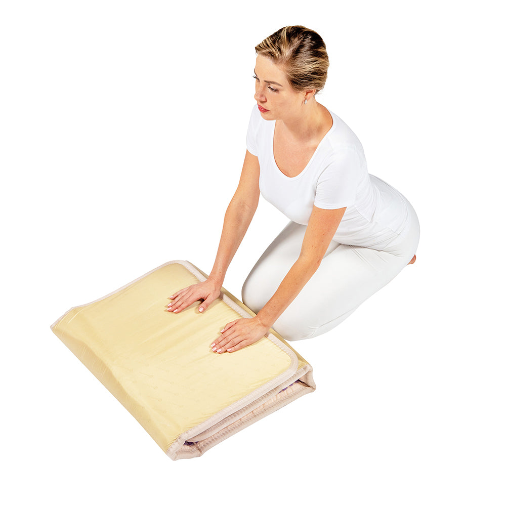 A person in a white outfit kneels on a plain white floor, folding a light beige blanket that provides comfort similar to the TAJ-Mat™ Full Short 6024 Firm - Photon PEMF InfraMat Pro® by HealthyLine.