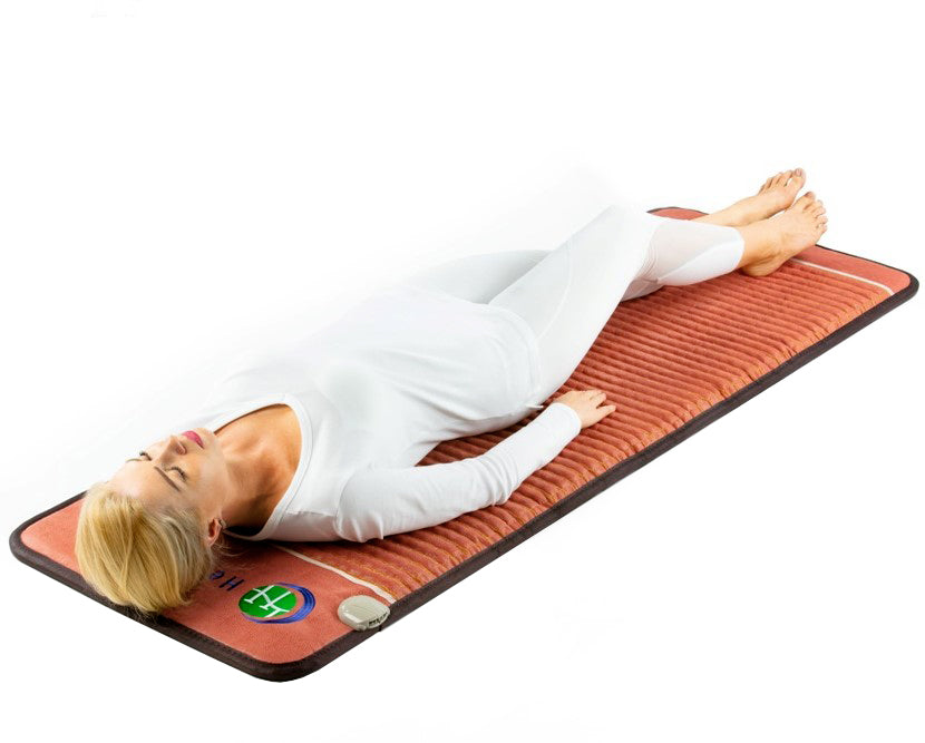 A person wearing white relaxes calmly on the HealthyLine TAO-Mat® Full 7224 Firm - PEMF InfraMat Pro®, a reddish-brown therapeutic mat with crushed gemstones. The attached control device indicates its advanced far infrared technology for relaxation or health benefits.