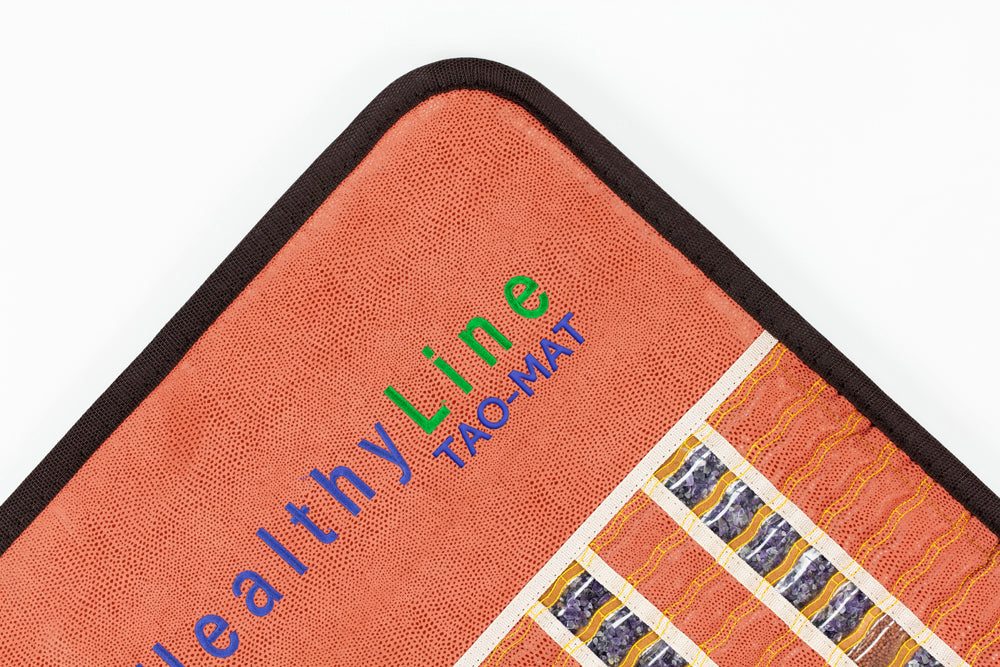 Close-up of a corner of a textured orange mat with black trim. The text "HealthyLine TAO-Mat® Full 7224 Firm - PEMF InfraMat Pro®" is embroidered in various colors. Enhanced with crushed gemstones, it features blue and white stripes and yellow wavy lines for elegance.