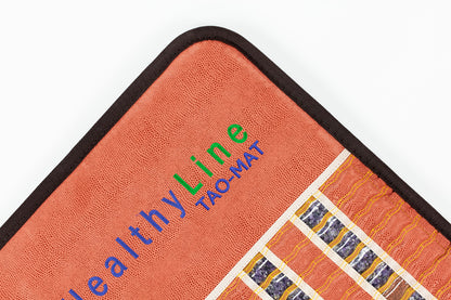 Close-up of a corner of a textured orange mat with black trim. The text "HealthyLine TAO-Mat® Full 7224 Firm - PEMF InfraMat Pro®" is embroidered in various colors. Enhanced with crushed gemstones, it features blue and white stripes and yellow wavy lines for elegance.