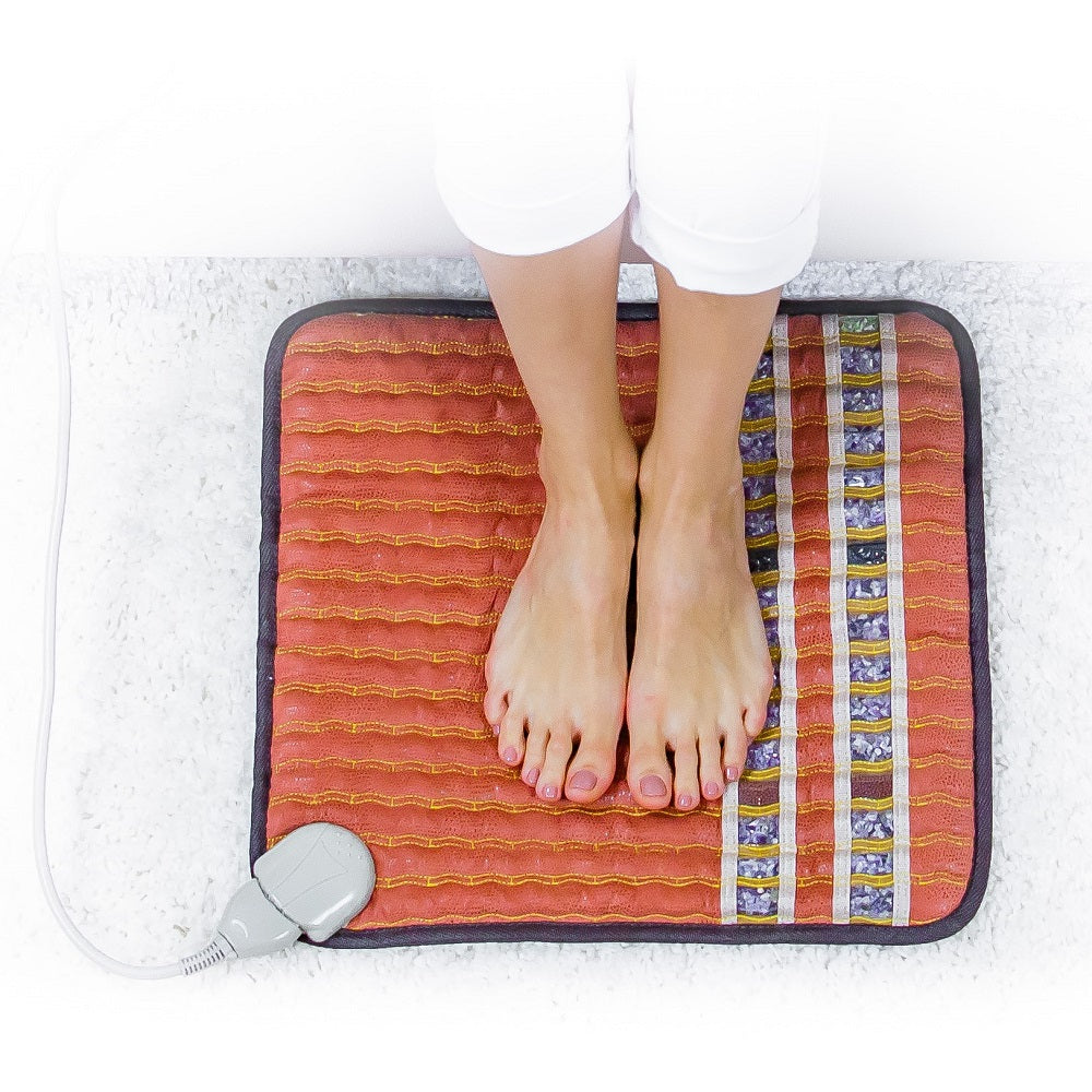 A person with bare feet rests on a HealthLine TAO-Mat® Small 1818 Soft InfraMat Pro®, featuring a striped design. This mat employs heat therapy and comes with a corded control, placed on a white carpet.