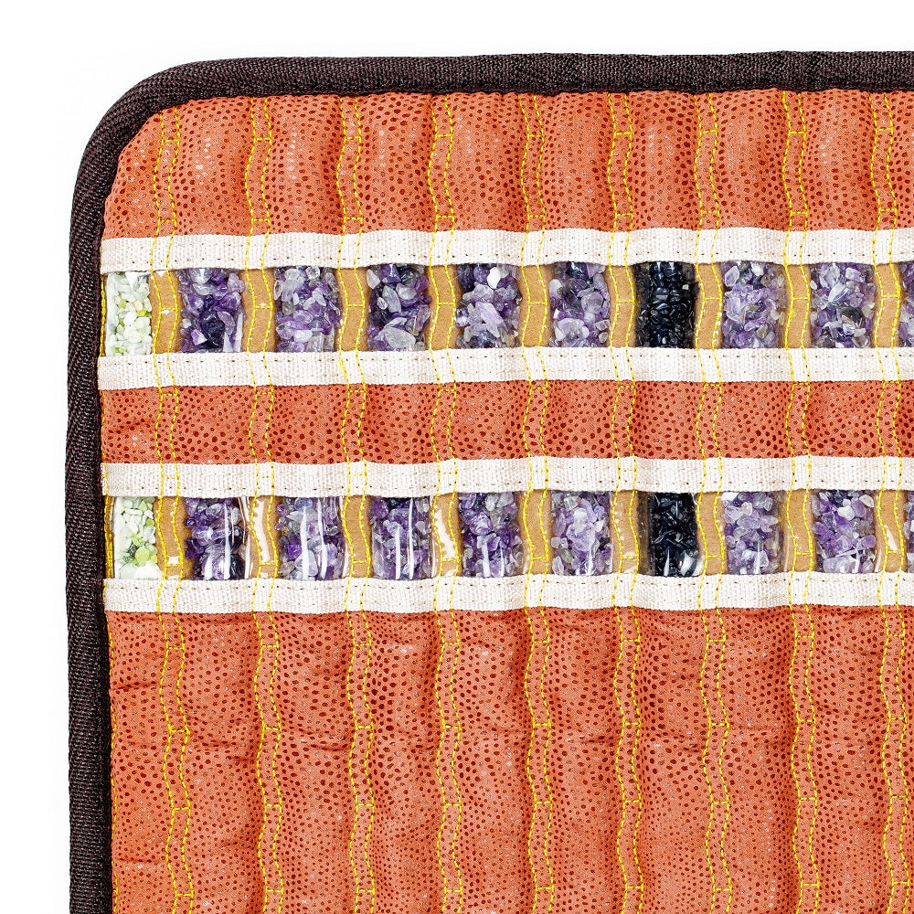 Close-up of a patterned textile showcasing horizontal rows of square segments filled with crushed natural gemstones, such as amethyst. The rows are divided by white and gold stitching on a textured orange fabric background with a dark edge, evoking the opulence of the HealthLine TAO-Mat® Small 1818 Soft InfraMat Pro®.