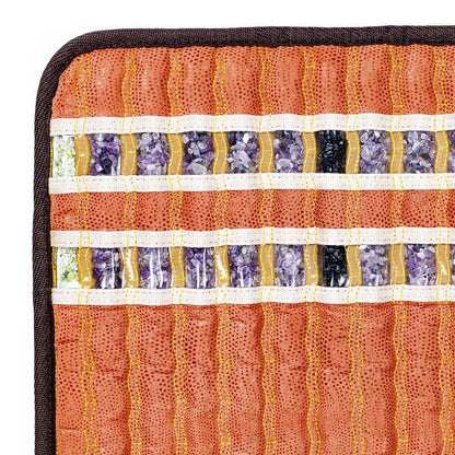 Close-up of a patterned textile showcasing horizontal rows of square segments filled with crushed natural gemstones, such as amethyst. The rows are divided by white and gold stitching on a textured orange fabric background with a dark edge, evoking the opulence of the HealthLine TAO-Mat® Small 1818 Soft InfraMat Pro®.