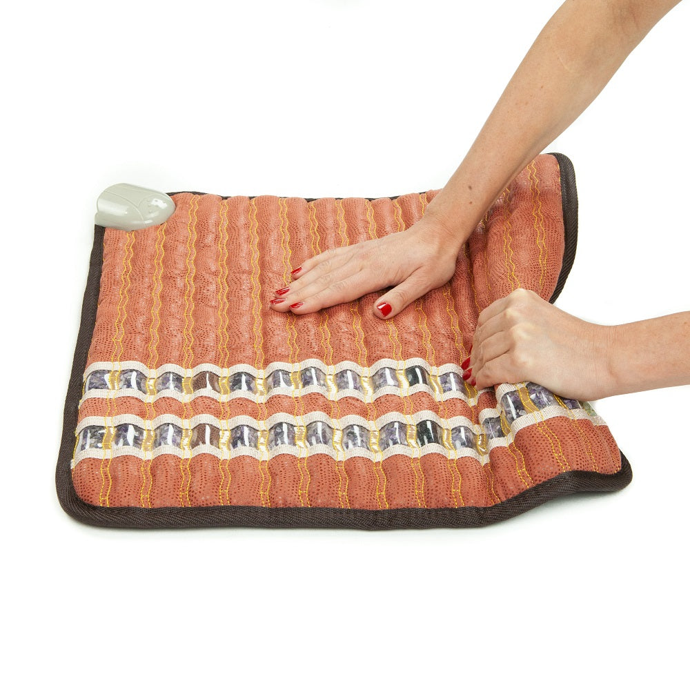 Hands adjusting a reddish-brown TAO-Mat® Small 1818 Soft InfraMat Pro® by HealthLine, which features black trim and white decorative patterns to enhance heat therapy. A white control switch is visible in the top left corner of the mat.
